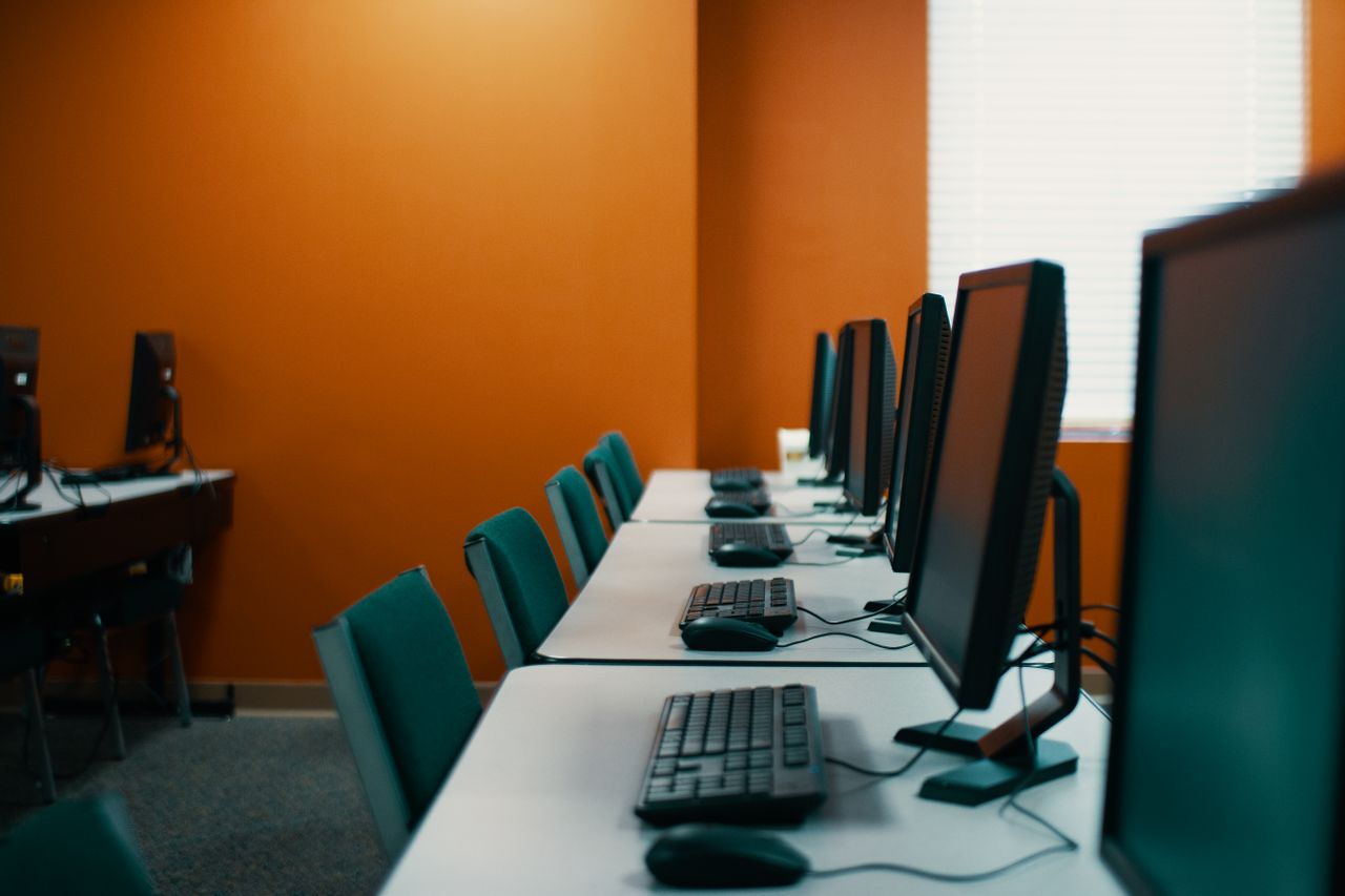 portfolio-03 Modern computers with keyboards and mice placed in row on table with chairs in contemporary workplace with orange walls